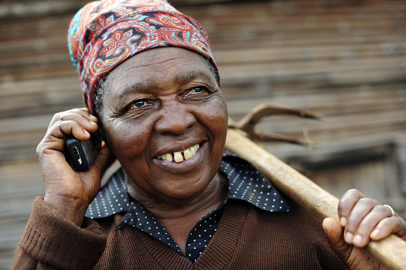 A female farmer speaks on phone, holding a phone with one hand and a farm tool in the other hand