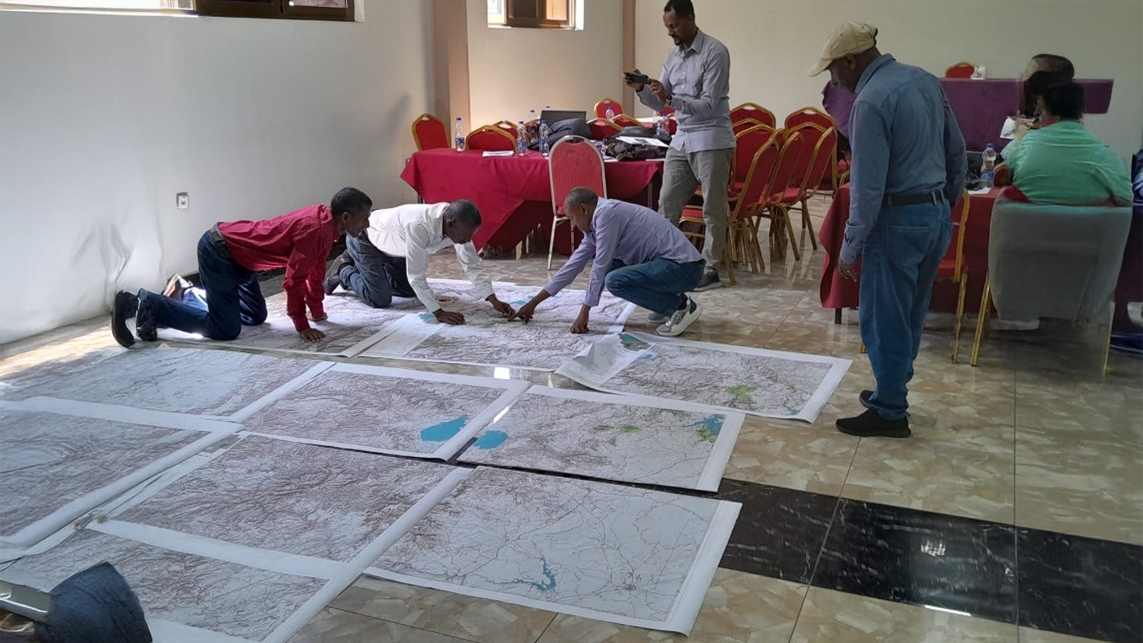 A group of people look over some large maps placed on the floor of a room