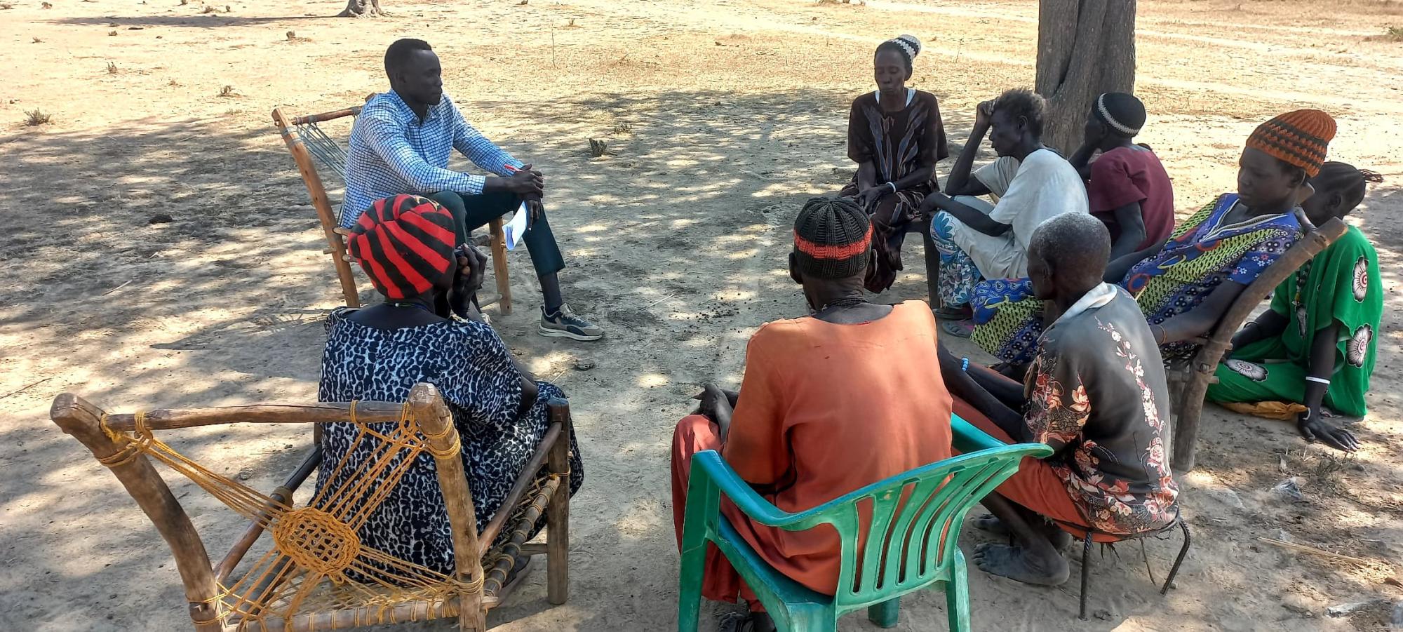 A group of people sit in a circle outside talking to a man with a notebook and pen in his hand
