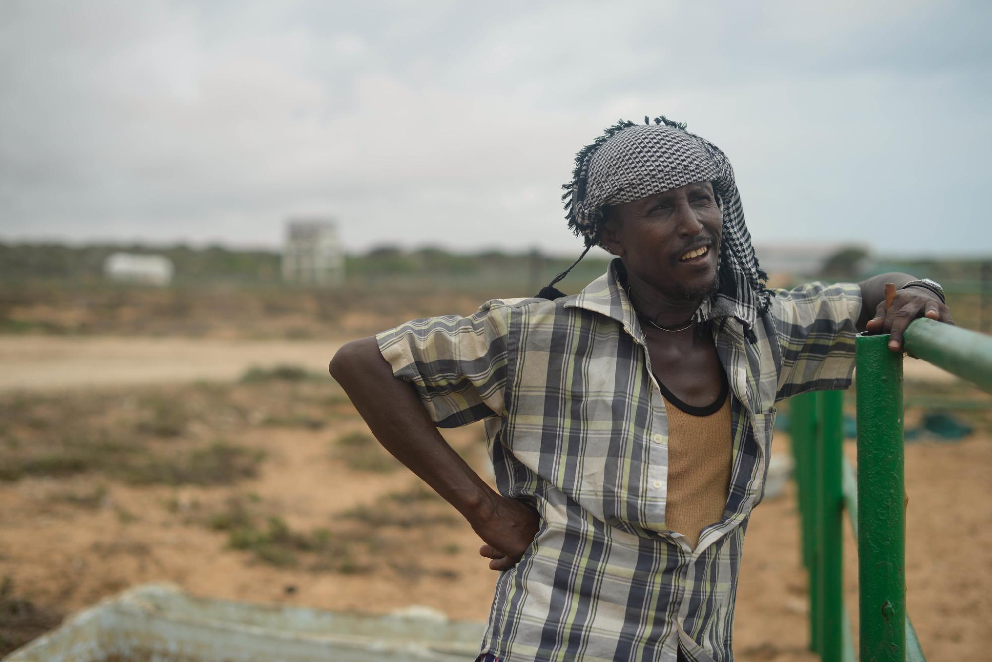 A man with a checked shirt leans against an iron fence with an arid landscape behind him