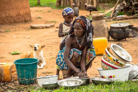 A woman bends down outside to wash dishes while a child stands behind her