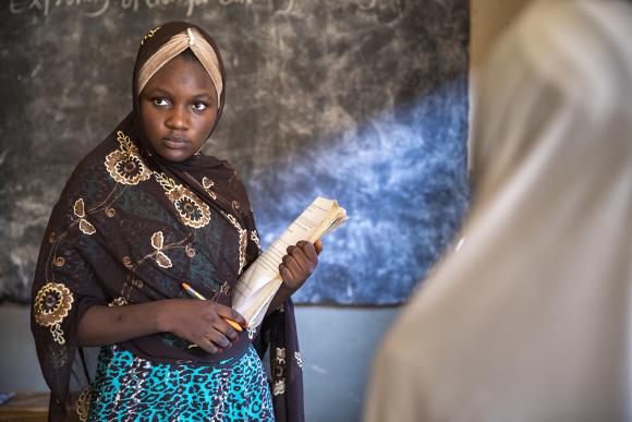 An adolescent girl stands in front of a chalkboard and holds a book