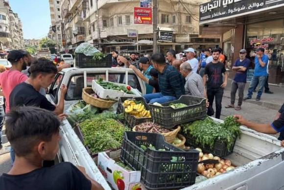 A group of men stand in a road near the back of a parked pickup trick filled with crates of vegetables and fruit