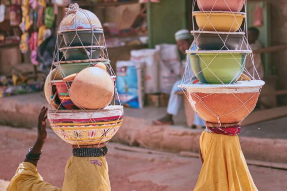 Two women carrying dishes stacked on their heads walk away from the camera