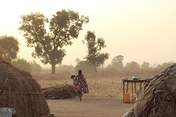 A woman stands in a Fulani camp with her back to camera and looks out to the trees
