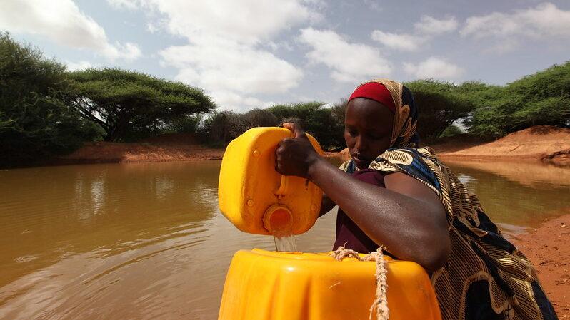 A woman stands at the edge of a reservoir filling large yellow water containers, with trees in background.