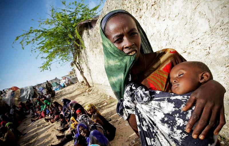 A woman stands next to a building holding a sleeping baby, with other women seated in the background.