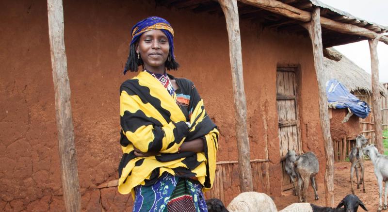 A young woman facing the camera stands outside in front of a mud building among goats