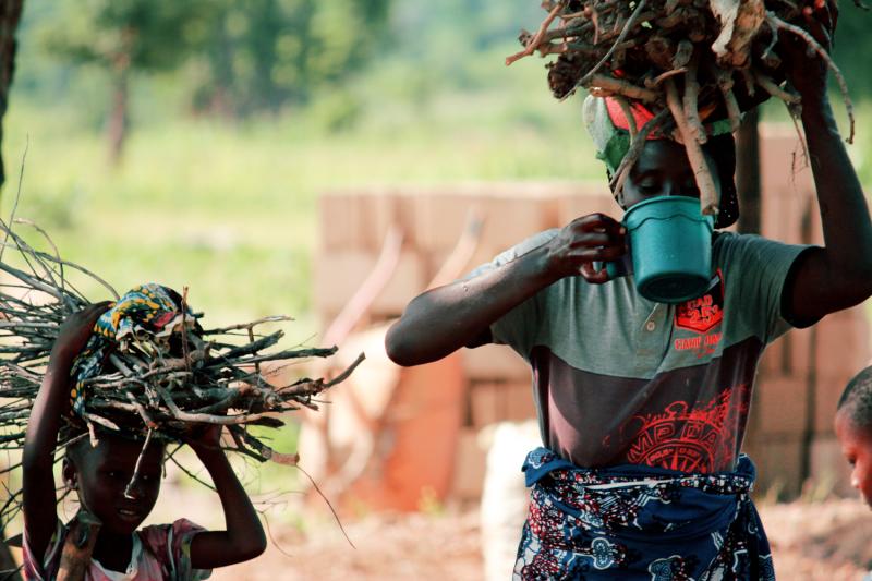 A woman and child stand outdoors holding branches on their heads. The woman is drinking from a blue plastic container.