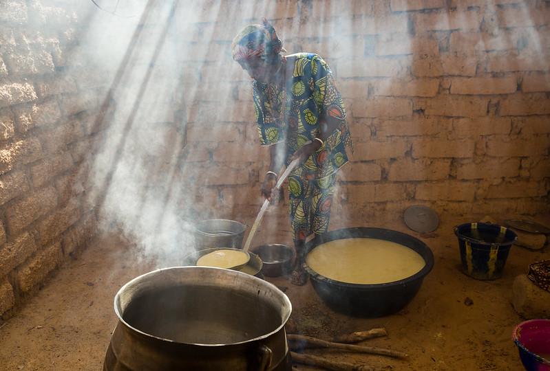 A woman stands inside building made from bricks in front of several large steaming pots, stirring their contents