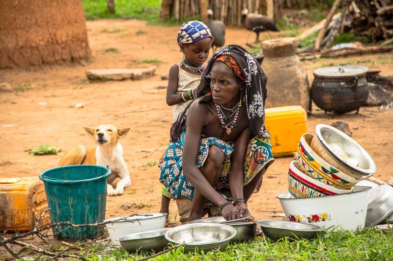 A woman crouches down on the ground washing dishes outside while a small girl stands behind her