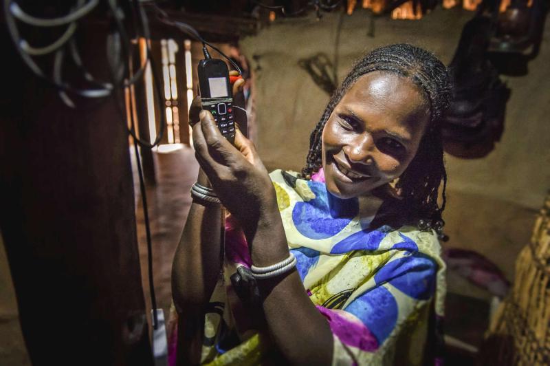 A woman indoors holding a mobile phone and smiling at the camera