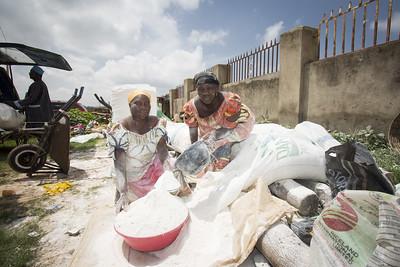 Two women sit among bags of cassava flour outdoors. One woman pours flour from a container into a large plastic bowl held by the other woman.
