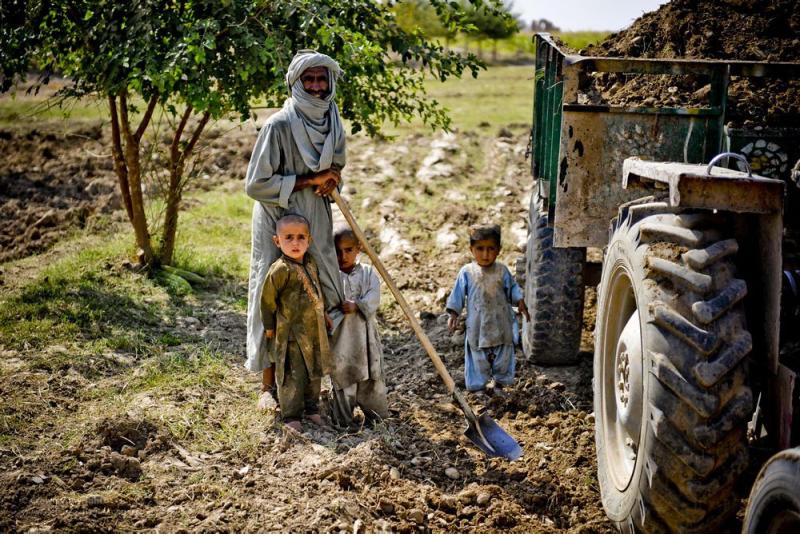 A man stands holding a shovel next to a tractor in a field, with three young children