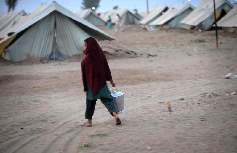 A young girl wearing a shawl over her head carries a bucket as she walls passed some tents