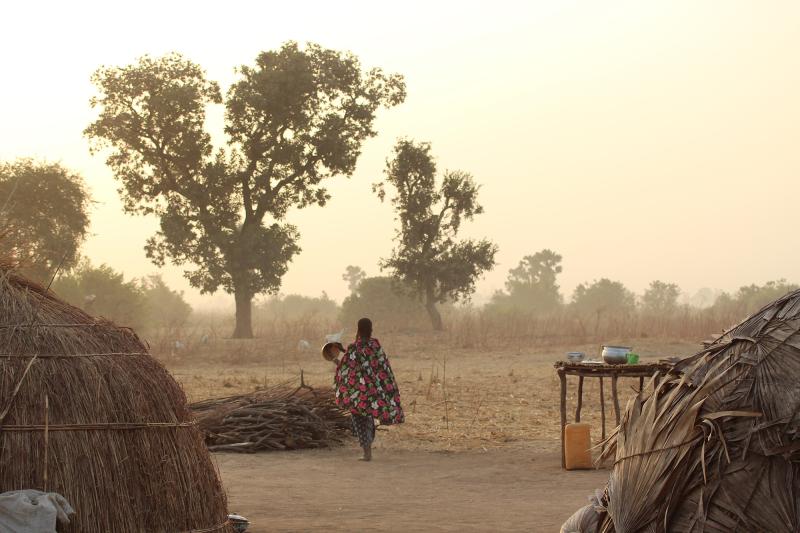 A woman stands in a Fulani camp with her back to camera and looks out to the trees