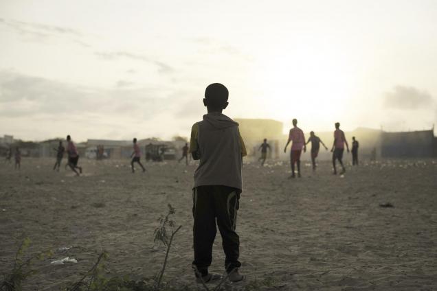 A young child looks on as older boys play football next to a camp for internally displaced persons (IDP) in Mogadishu, Somalia.