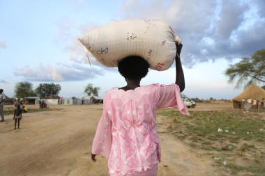 The back of a woman wearing pink as she walks away from the camera carrying a sack of provisions on her head