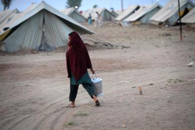 A young girl wearing a shawl over her head carries a bucket as she walls passed some tents