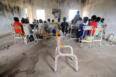  A group of children sit in a rundown classroom looking at the blackboard with their backs to the camera