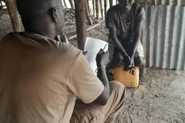 A pastoralist sits in the background facing the camera while being interviewed