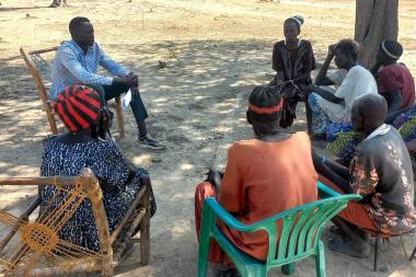 A group of people sit in a circle outside talking to a man with a notebook and pen in his hand
