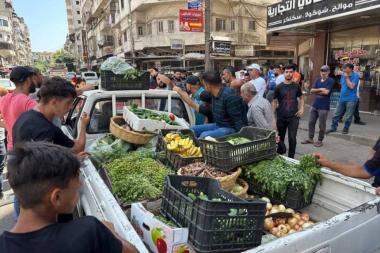 A group of men stand in a road near the back of a parked pickup trick filled with crates of vegetables and fruit