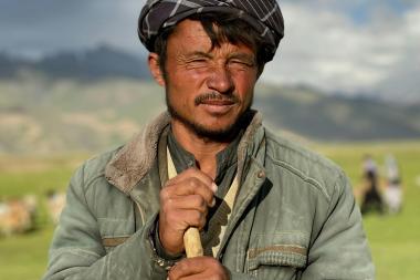 Close up of a shepherd holding a stick and looking at the camera with a blurred background of grass and livestock