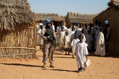 A soldier in combat fatigues stand holding a gun in a dusty camp while some children in white clothes walk past
