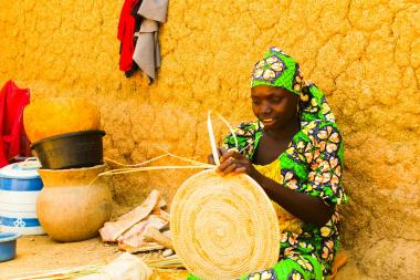 A woman wearing brightly-coloured clothes leans against a wall outside and weaves with straw