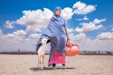 A woman standing with a goat in front of a blue sky dotted with white clouds